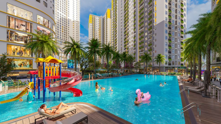 Hình ảnh phối cảnh khu tiện ích ngoài trời với hồ bơi rộng, khu vui chơi dưới nước dành cho trẻ em, ghế nghỉ thư giãn và hàng cây xanh bao quanh. Các tòa căn hộ cao tầng cùng khối đế thương mại tạo nên không gian sinh hoạt kết hợp giữa vận động, nghỉ ngơi và vui chơi cho gia đình.