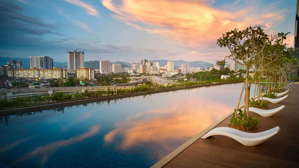 Hình ảnh hồ bơi ngoài trời với mặt nước phản chiếu ánh hoàng hôn, bên cạnh là lối đi lát sàn gỗ, ghế thư giãn và hàng cây xanh. Phía xa là cảnh quan đô thị với các tòa nhà cao tầng và tuyến đường giao thông. Hình ảnh phù hợp minh họa cho nội dung về tiện ích hồ bơi, không gian thư giãn và chất lượng sống tại dự án căn hộ.